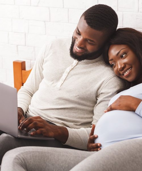 Lovely pregnant afro couple using laptop while resting at home, laying on bed, woman leaning on man shoulders, copy space